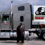 Trailer Transit Inc. | A man stands beside a large gray semi-truck. A banner in the image reads "Owner Operator of the Month - Trailer Transit Inc." with a truck graphic. Trees and other trucks are in the background.