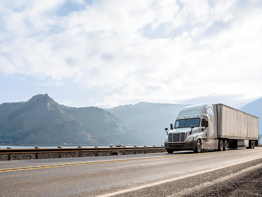 Trailer Transit Inc. | A large semi-truck with a trailer, likely operated by owner-operator transport companies, drives on a highway next to mountains and a body of water under a partly cloudy sky.