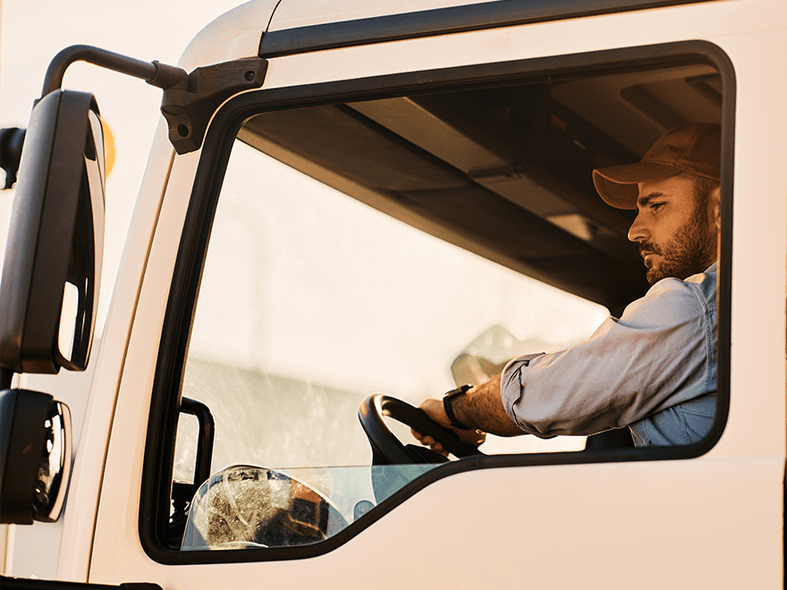 Trailer Transit Inc. | A man wearing a cap is driving a truck, viewed from the passenger side window, with sunlight filtering through. The scene captures the daily life of owner-operators working for transport companies.