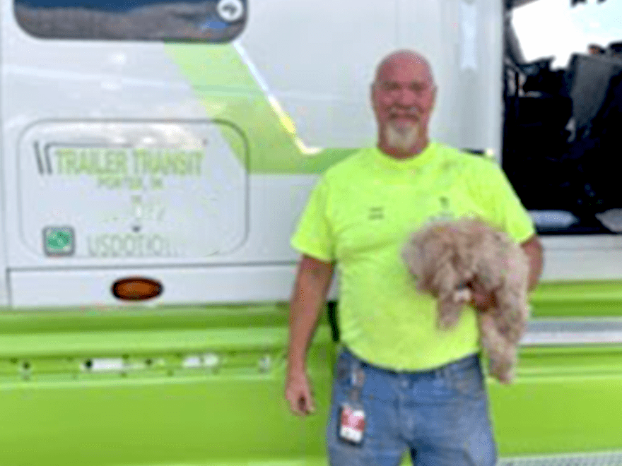 Trailer Transit Inc. | A man wearing a neon yellow shirt stands in front of a truck, holding a small dog. The truck has “TRAILER TRANSIT” printed on it along with other text and markings, indicating it's part of an owner-operator transport company.