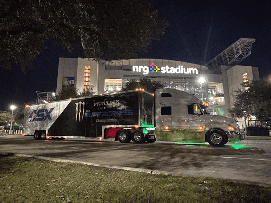 Trailer Transit Inc. | A semi-truck with "Monster Energy Supercross" branding, owned by one of the top owner operator transport companies, is parked outside the illuminated NRG Stadium at night.