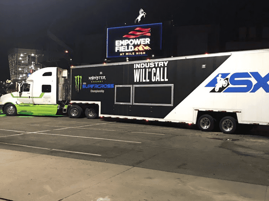 Trailer Transit Inc. | A large semi-trailer truck, often used by owner operator transport companies, is parked at night in front of Empower Field. The trailer boasts "Monster Energy Supercross" and "Industry Will Call" signage.