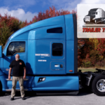 Trailer Transit Inc. | A man stands proudly in front of a blue semi-truck under a "Owner Operator Trailer Transit of the Month" banner, against a backdrop of autumn-colored trees.