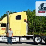 Trailer Transit Inc. | Man standing next to a large yellow semi-truck with "Trailer Transit Inc." branding. A logo in the corner reads, "Owner Operator of the Month, Trailer Transit Inc." Trees are visible in the background, showcasing the pride of owner operators who work with Trailer Transit.