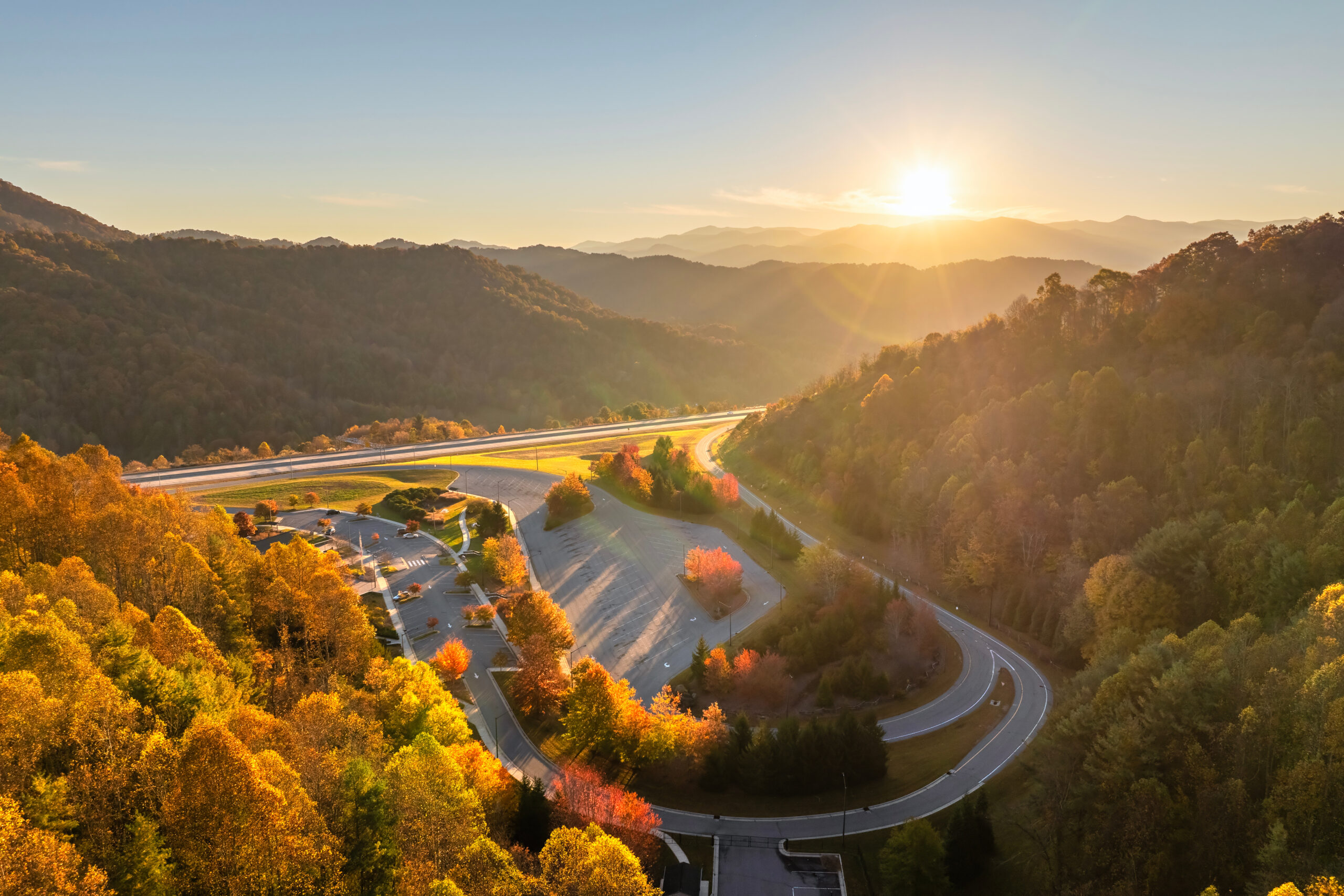 Trailer Transit Inc. | A winding road curves through a forested mountainous area at sunset, with vibrant autumn foliage and a sunlit sky.