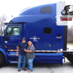 Trailer Transit Inc. | December Owner Operators of the Month, Larry & Barbara, proudly stand in front of their blue semi-truck adorned with the "Trailer Transit Inc." logo.